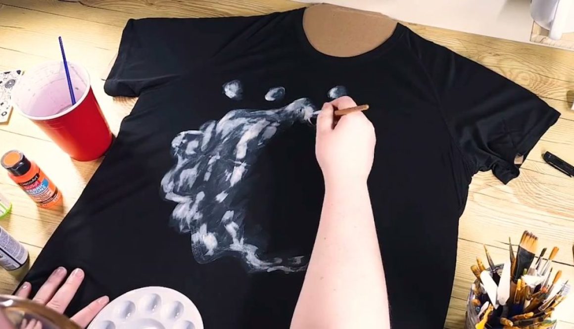 # Description A person is painting white highlights and details onto a black t-shirt design of a wolf's head using a brush, with painting supplies and a palette visible on the wooden work surface.