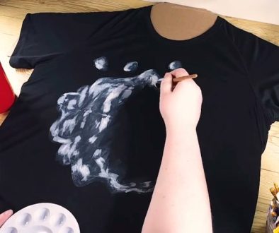 # Description A person is painting white highlights and details onto a black t-shirt design of a wolf's head using a brush, with painting supplies and a palette visible on the wooden work surface.