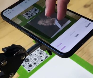 # Image Description A person is using the Da Vinci Eye app on their smartphone to trace or draw a portrait by positioning the phone's camera over a reference image on a green drawing board, with editing options like "Opacity," "Hide," and "More" visible on the screen.