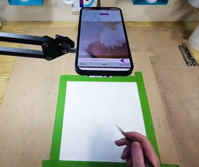 # Description A person is using the Da Vinci Eye app on a smartphone to trace or draw a hand on paper with a green border, while the phone displays a real-time video feed to guide their drawing.