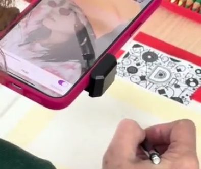 # Drawing Tutorial Description A person is using a smartphone mounted on a stand to project an image as a reference while sketching with a pencil on paper, surrounded by colorful art supplies including colored pencils.